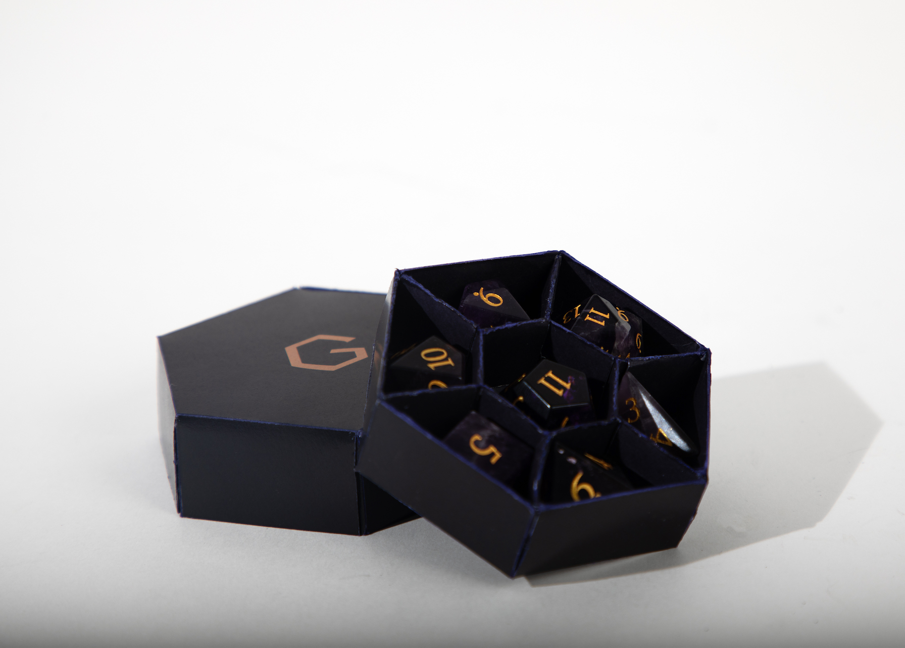 Die sechseeckige violette Box ist geöffnet zu sehen und lehnt auf ihrem Deckel. Innen sind sieben Fächer mit amethysternen Würfeln, wobei sechs der Fächer trapezförmig im Kreis um das siebte in der Mitte angeordnet sind.